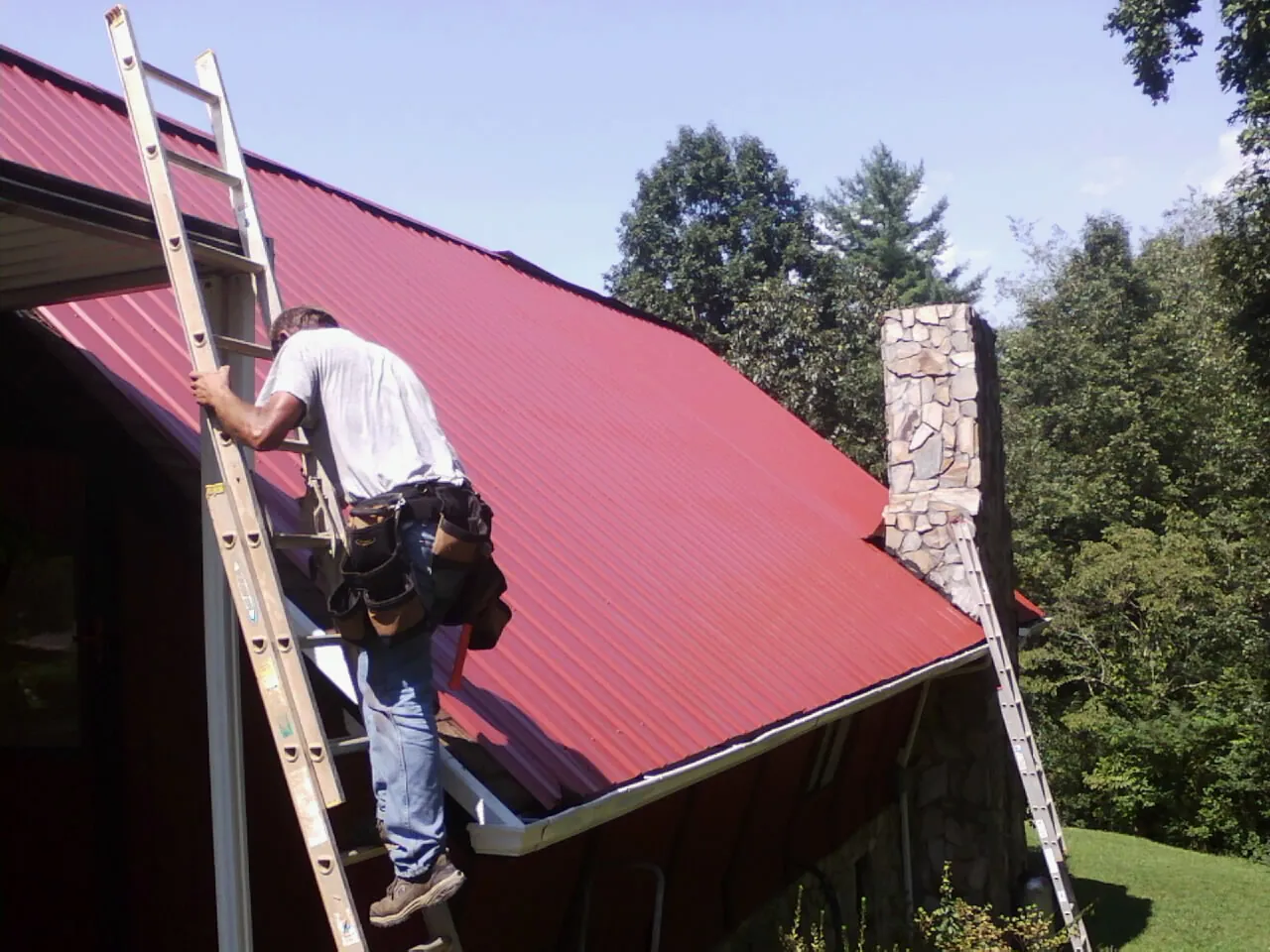 Roofer on a ladder beside a red metal roof and stone chimney — Soffit & Fascia Repair by Roofing Brothers in Bryn Athyn